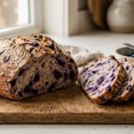 A large round loaf of blueberry sourdough bread on a wooden cutting board, with several slices cut.